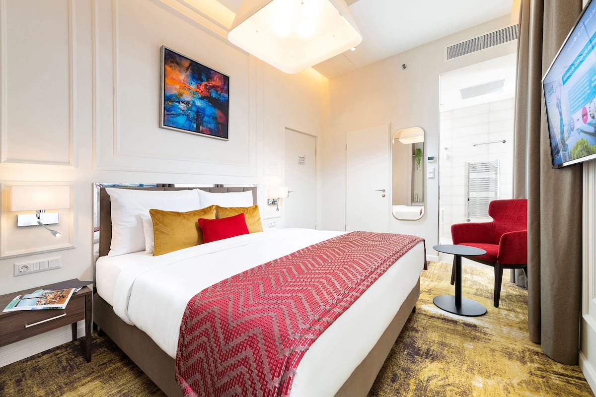 Ein Hotelzimmer mit einem großen Bett, einem roten Sessel, einem kleinen Tisch und einem Fernseher an der Wand.