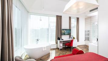 Habitación de hotel moderna con un ventanal grande, bañera, escritorio con silla y cama con ropa de cama roja.