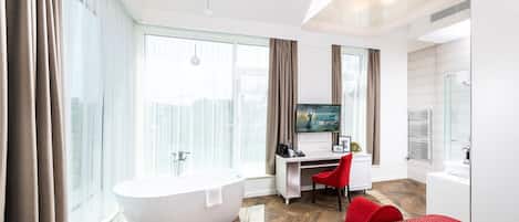 A modern hotel room with a large window, a bathtub, a desk with a chair, and a bed with red bedding.