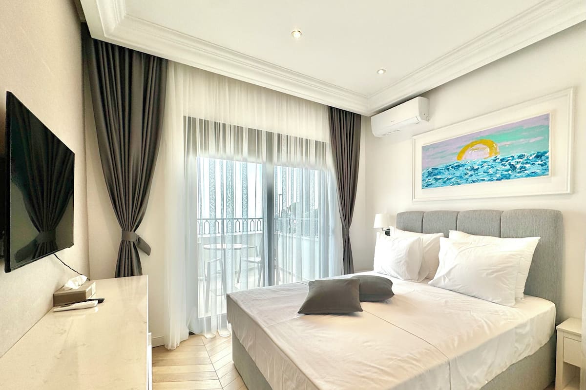 A hotel room with a bed, a flat-screen TV, a painting of a beach scene, and a balcony with a view.