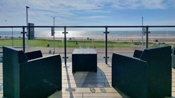 An outdoor seating area with two dark-colored sofas and a table, overlooking a beach and the sea.