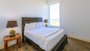 Une chambre avec une grande fenêtre, un lit recouvert de draps blancs et une table de chevet avec une lampe.