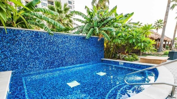 Une piscine ornée de motifs en mosaïque, entourée de plantes tropicales et d’un palmier.