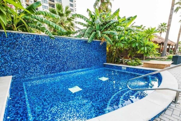 Una piscina con diseño de mosaicos, rodeada de plantas tropicales y una palmera.
