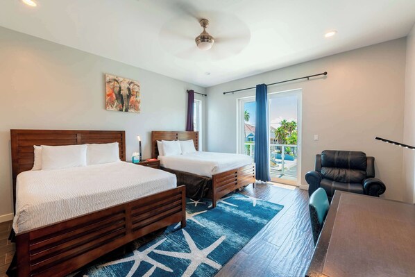 Habitación de hotel con dos camas, una mesa de comedor, un sillón de cuero y un balcón con vista a palmeras.