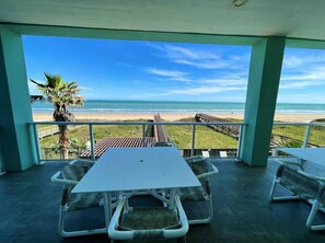 Zona exterior coberta amb taula i cadires, amb vista a una platja que inclou una palmera i una terrassa de fusta.