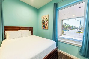 A bedroom with a bed, a window with a view of the street, and a painting on the wall.