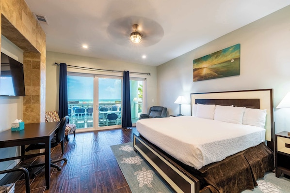 A hotel room with a large bed, a desk, a chair, a TV, and a view of the ocean through the window.