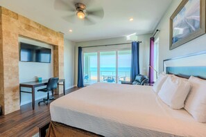 A hotel room with a large bed, a desk, a chair, a TV mounted on a stone wall, and a view of the sea through large windows.
