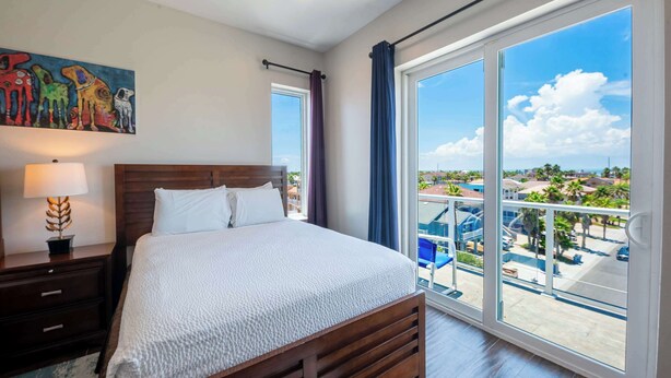 A hotel room with a large bed, a nightstand with a lamp, a painting on the wall, and a balcony with a pool view. - Dolphin Paradise  (South Padre Island)