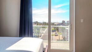 A hotel room with a bed, a view of a residential area through a sliding glass door, and a balcony with a railing. - Dolphin Paradise  (South Padre Island)