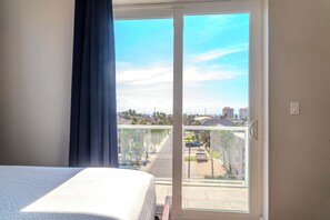 A hotel room with a bed, a view of a residential area through a sliding glass door, and a balcony with a railing.