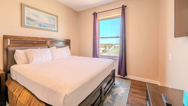 A neatly made bed with a wooden headboard, a painting on the wall, a window with purple curtains, and a remote control on the nightstand. - Dolphin Paradise  (South Padre Island)