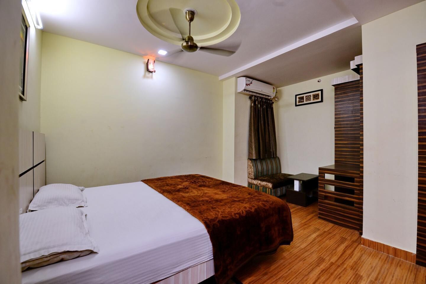 A hotel room with a bed, a ceiling fan, an air conditioning unit, a small table, and a seating area with a sofa and curtains.