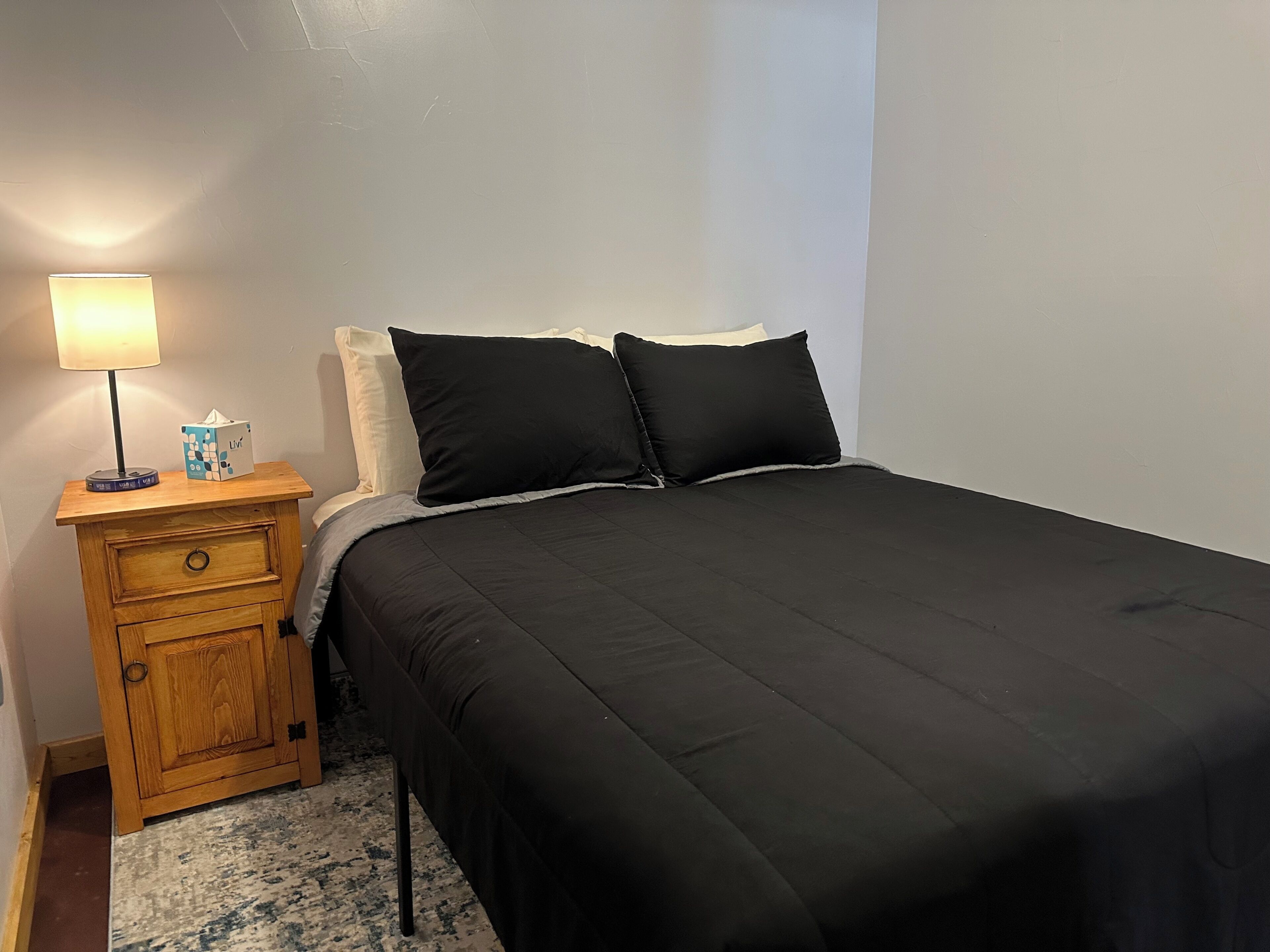 Ein Bett mit schwarzer Bettwäsche, ein Nachttisch aus Holz mit einer Lampe und einer Taschentuchbox.