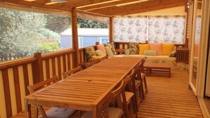 A wooden deck with a table and chairs, a sofa with cushions, and a view of the outdoors through large windows. - 6-seater mobile home, large covered terrace, in a 4-star campsite. (Le Muy)