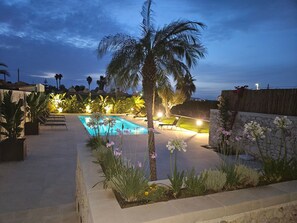 Une piscine entourée de plantes et de palmiers, avec des chaises longues et un mur en pierre.