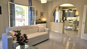 A modern living room with a sofa, a glass coffee table, and a potted plant. There is a kitchen area visible through an archway, featuring a dining table and chairs. - Villa Merilusi by Ortigiaapartments (Syracuse)