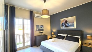 A modern bedroom with a large bed, a nightstand, a dresser, and a window with curtains. - Villa Merilusi by Ortigiaapartments (Syracuse)
