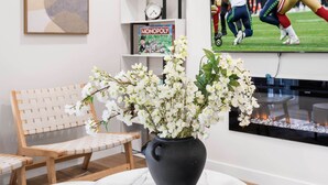A modern living room with a television displaying a football game, a white sofa, a small round table, and a vase with white flowers.