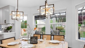A dining area with a large table set for six, surrounded by wicker chairs. There are pendant lights above the table, and the room has large windows offering a view of the outdoors.