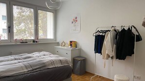 Camera da letto con un letto, una cassettiera, un termosifone, una finestra con persiane, una lampada a sospensione, un appendiabiti e uno sgabello.