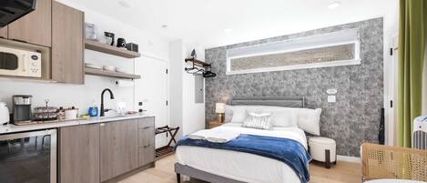 Ein modernes Hotelzimmer mit Küchenzeile, einem Bett mit grauem Kopfteil, einem Nachttisch, einer Lampe und einem Fenster mit Jalousien.