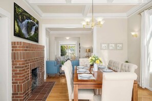 A dining area with a brick fireplace, a wooden dining table set for four, blue and white sofas, and a chandelier.