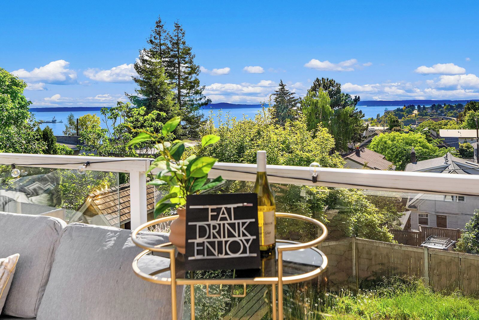 A balcony with a view of a body of water, a bottle of wine, and a sign that reads 'FAT DRINK ENJOY'.