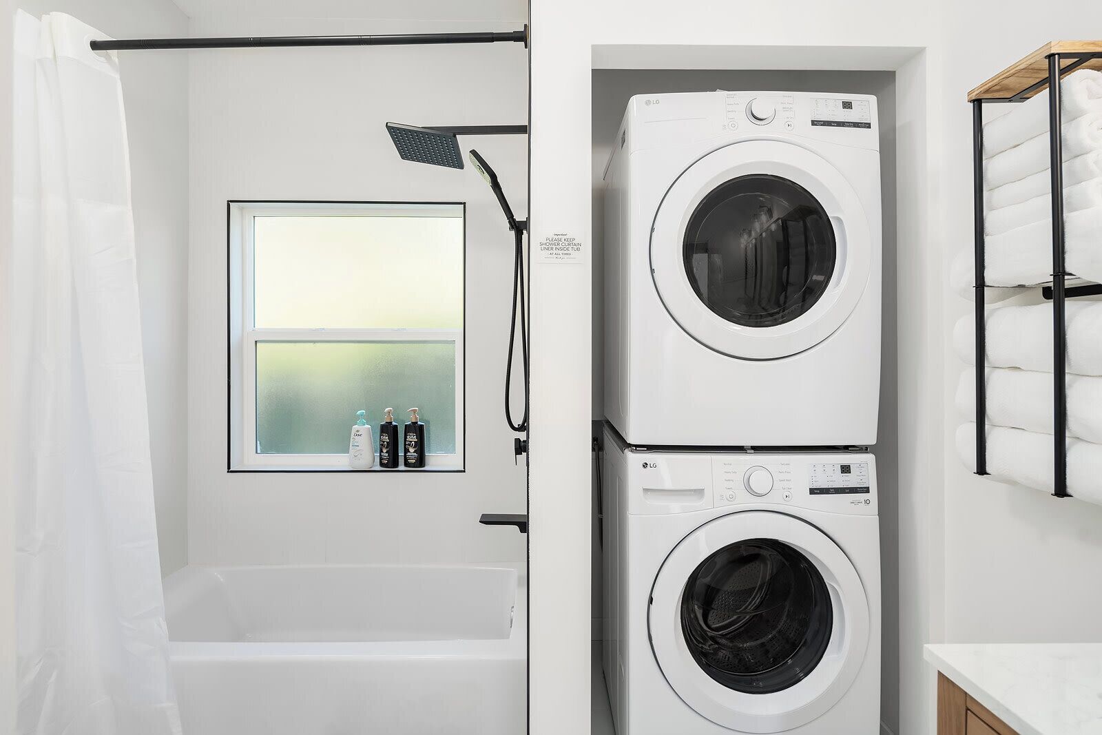 A laundry room with two stacked washing machines, a window with bottles, and a shower curtain.
