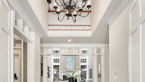 A spacious foyer with a grand chandelier, a round table with a vase of flowers, and a view of the outdoors through large windows.
