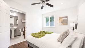 A bedroom with a bed, a ceiling fan, a window, and a bathroom visible through an open door.