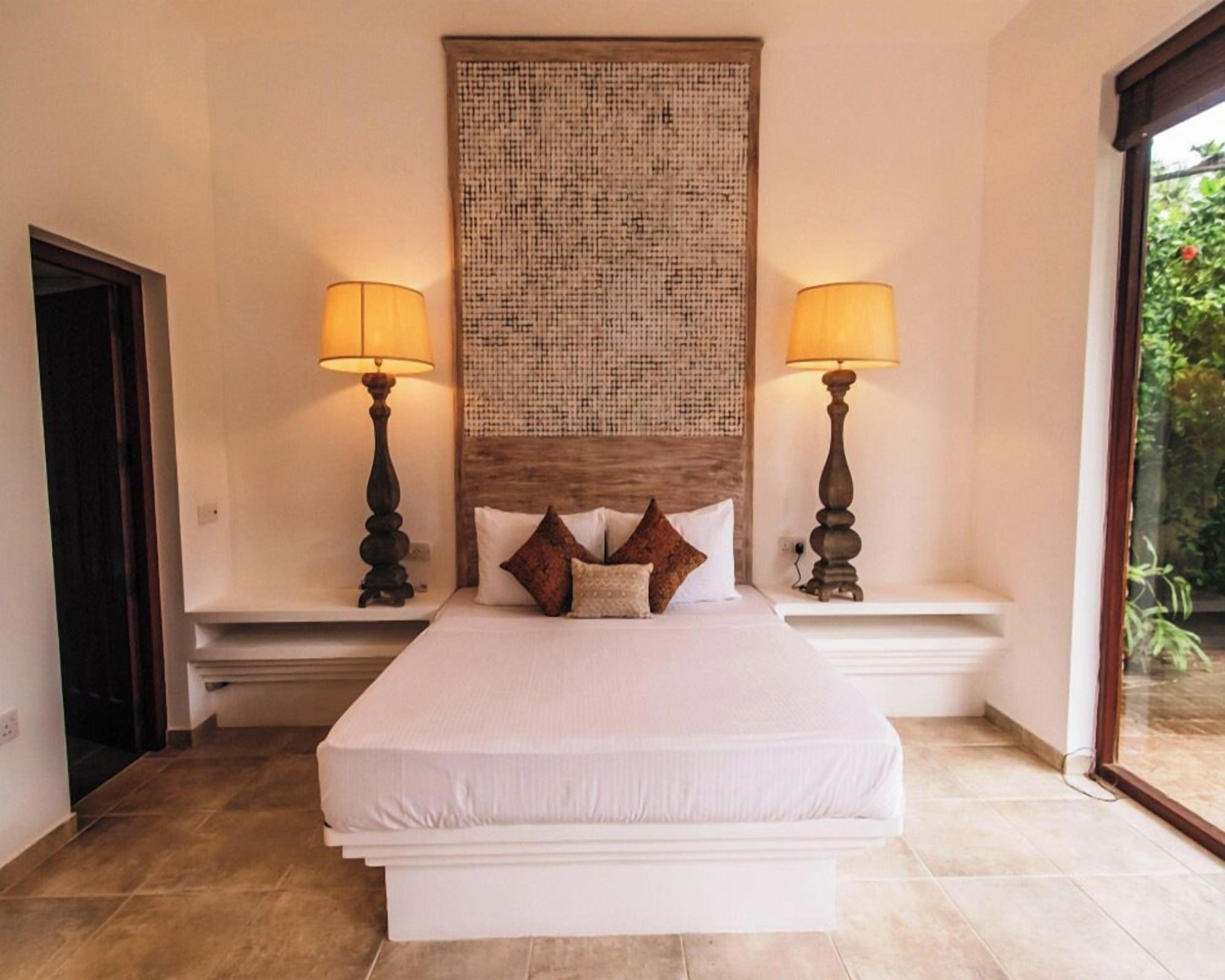 A large bed with a headboard, two bedside lamps, and a view of greenery through a window.