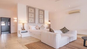 Ein modernes Wohnzimmer mit einer Couch, einem Couchtisch und einem Bett mit Kopfteil. Eine Tür führt in einen weiteren Raum, und an der Wand ist eine Klimaanlage angebracht.