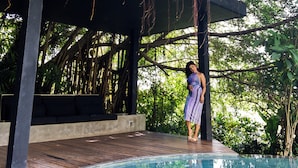 Un espace piscine doté d’une terrasse couverte, entouré d’une végétation luxuriante et d’arbres.