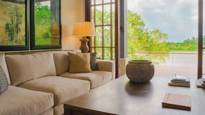 Une pièce avec un canapé beige, une table avec une lampe, un vase et des livres. Il y a une fenêtre donnant sur un paysage verdoyant.