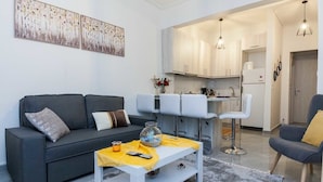 A modern living room with a grey sofa, white coffee table, and a kitchen area with white cabinets and bar stools.
