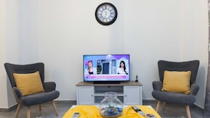 A living room with a television displaying a news program, two armchairs with cushions, a coffee table with a glass jar and remote controls, and a wall clock.