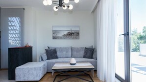 A modern living room with a sectional sofa, a wooden coffee table, a black metal frame, a ceiling light fixture, a painting, and a view through a large window.