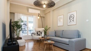 Een moderne woonkamer met een grijze bank, een witte fauteuil en een houten salontafel. Er staan twee potplanten, een zwarte kast en een groot raam met gordijnen.