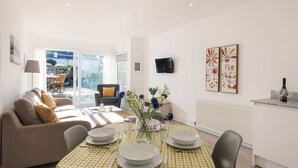 A modern living room with a sofa, dining table set for four, and a view of the garden through a glass door.