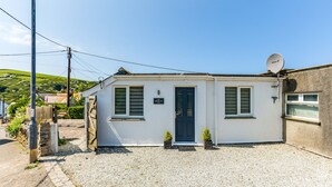 A small, single-story building with a dark door, two windows with shutters, a satellite dish, and a gravel driveway.