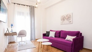 A modern living room with a purple sofa, two small white tables, a white desk with a chair, and a large window with curtains. - Sleek & Modern Apartment Near Metaxourgeio (Athens)