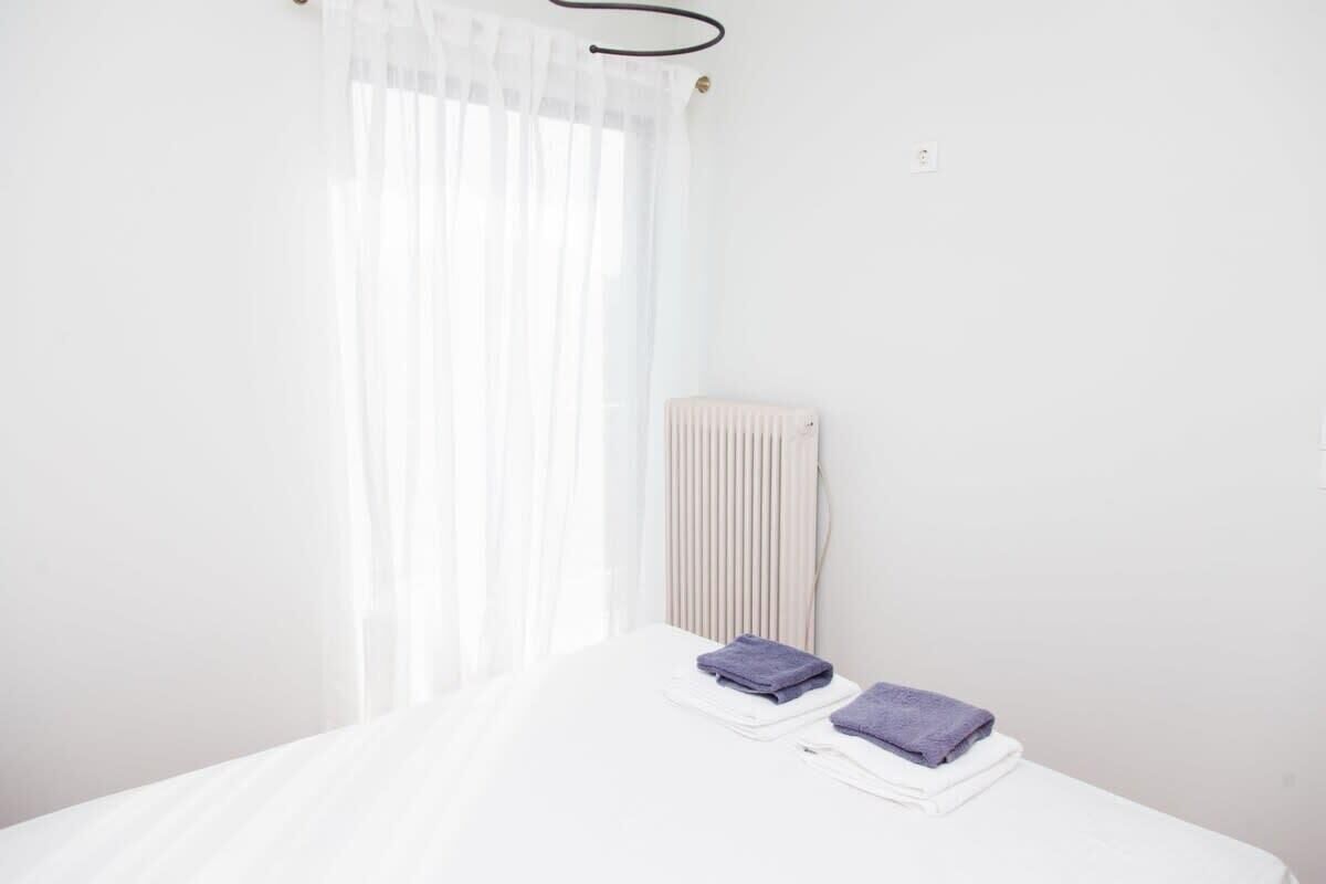 A clean, white room with a bed, a radiator, and a window with sheer curtains.