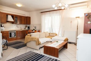 A modern kitchen with wooden cabinets, a dining area with a sofa, and a large window with sheer curtains. - Cozy modern apt in Peristeri (Athens)