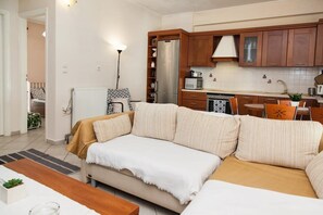 A modern living room with a sofa, a dining table, and a kitchen area with wooden cabinets and appliances. - Cozy modern apt in Peristeri (Athens)
