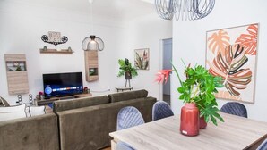 Un salon moderne avec un canapé gris, une table à manger en bois et un téléviseur. On trouve des plantes en pot sur la table et une étagère murale ornée d’objets décoratifs.