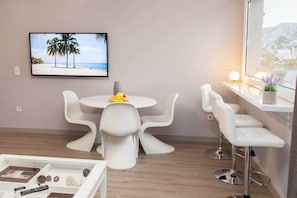 Une pièce moderne avec une table ronde, des chaises blanches, un téléviseur fixé au mur et une fenêtre donnant sur un beau paysage.