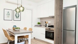 Uma cozinha moderna com armários brancos, uma geladeira de aço inoxidável, uma área de jantar com mesa e cadeiras, e luminárias pendentes.