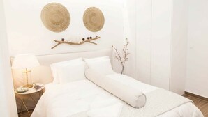 Uma cama bem arrumada com lençóis brancos, uma mesa de cabeceira com abajur e objetos decorativos, e dois painéis de parede trançados.